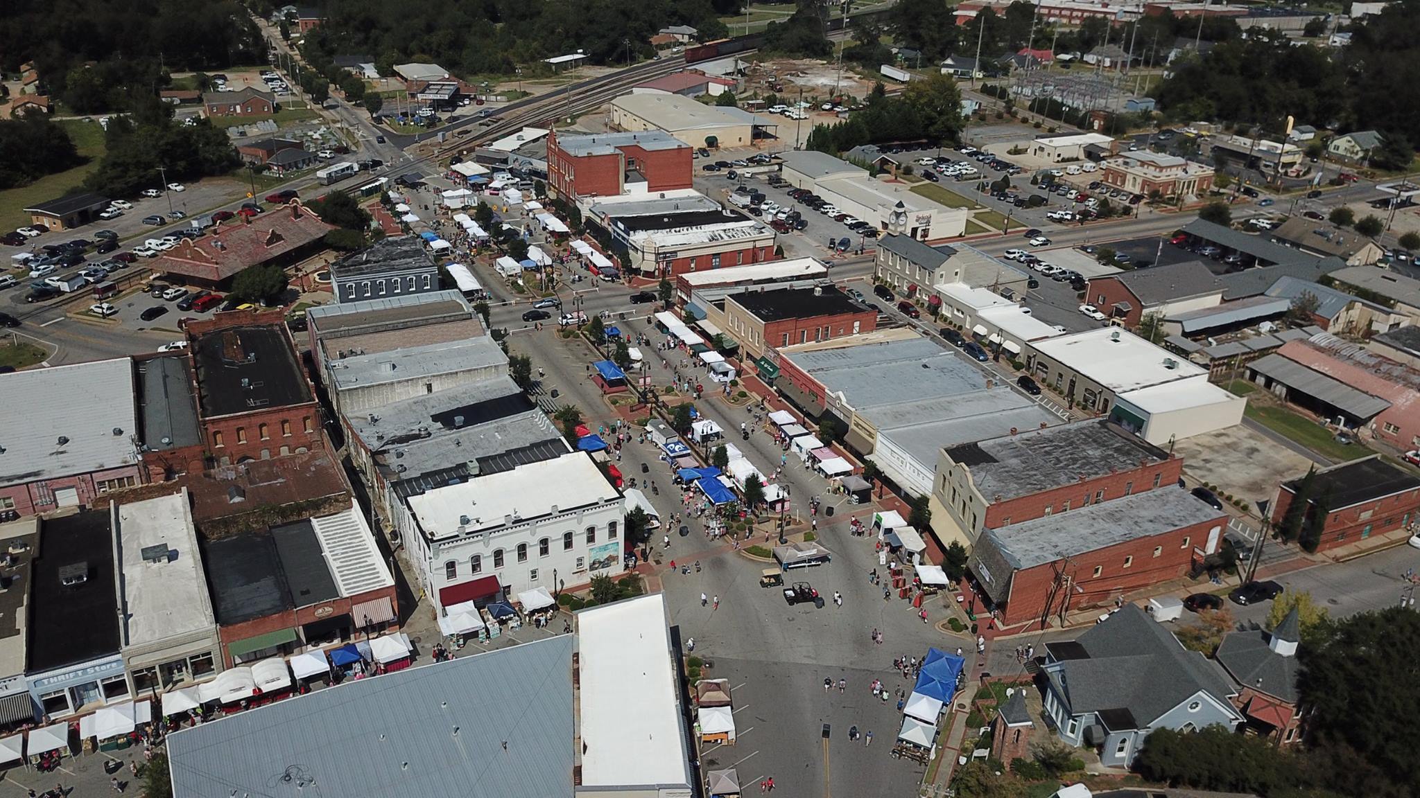 Aerial view of a busy outdoor market with stalls and people on a street, surrounded by buildings and trees.