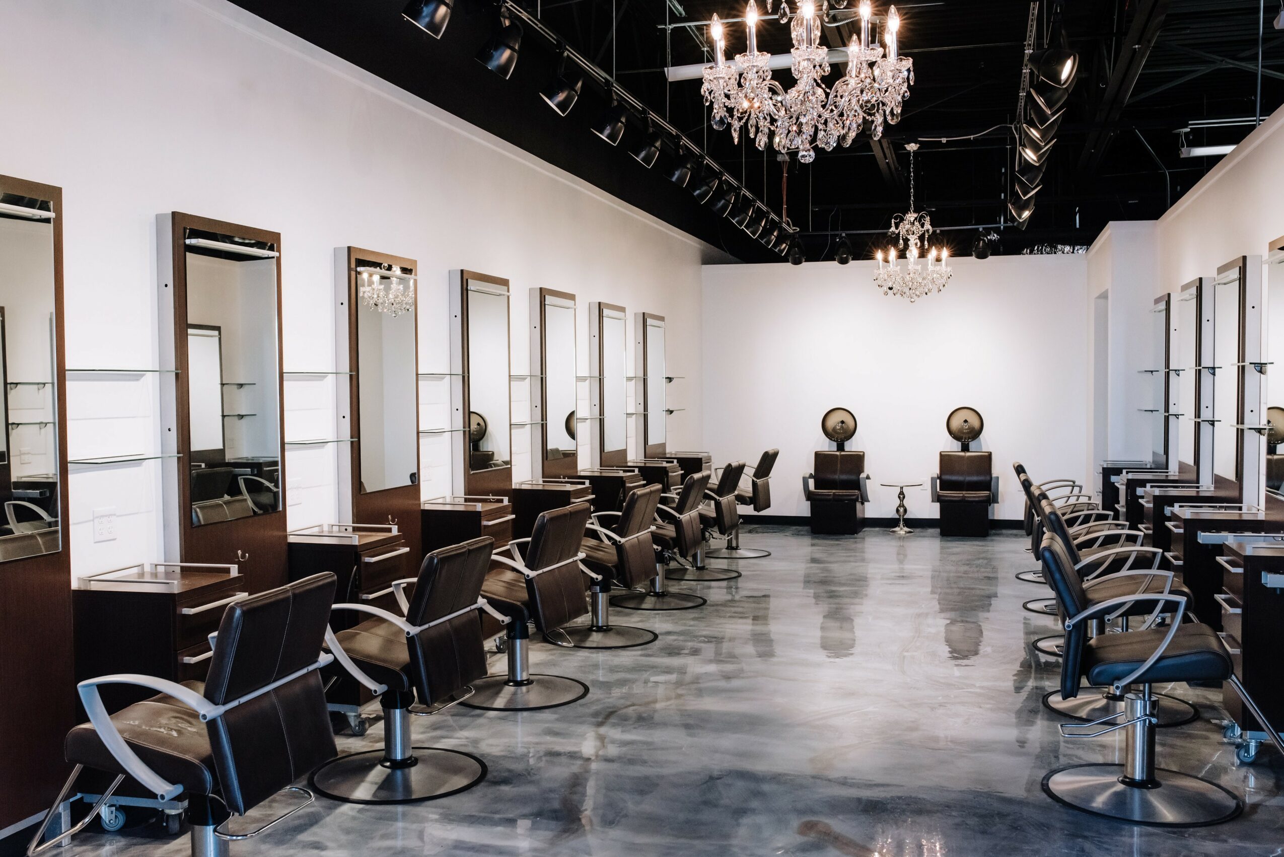 Empty salon with styling chairs, mirrors, chandeliers, and a polished floor, ready for clients.