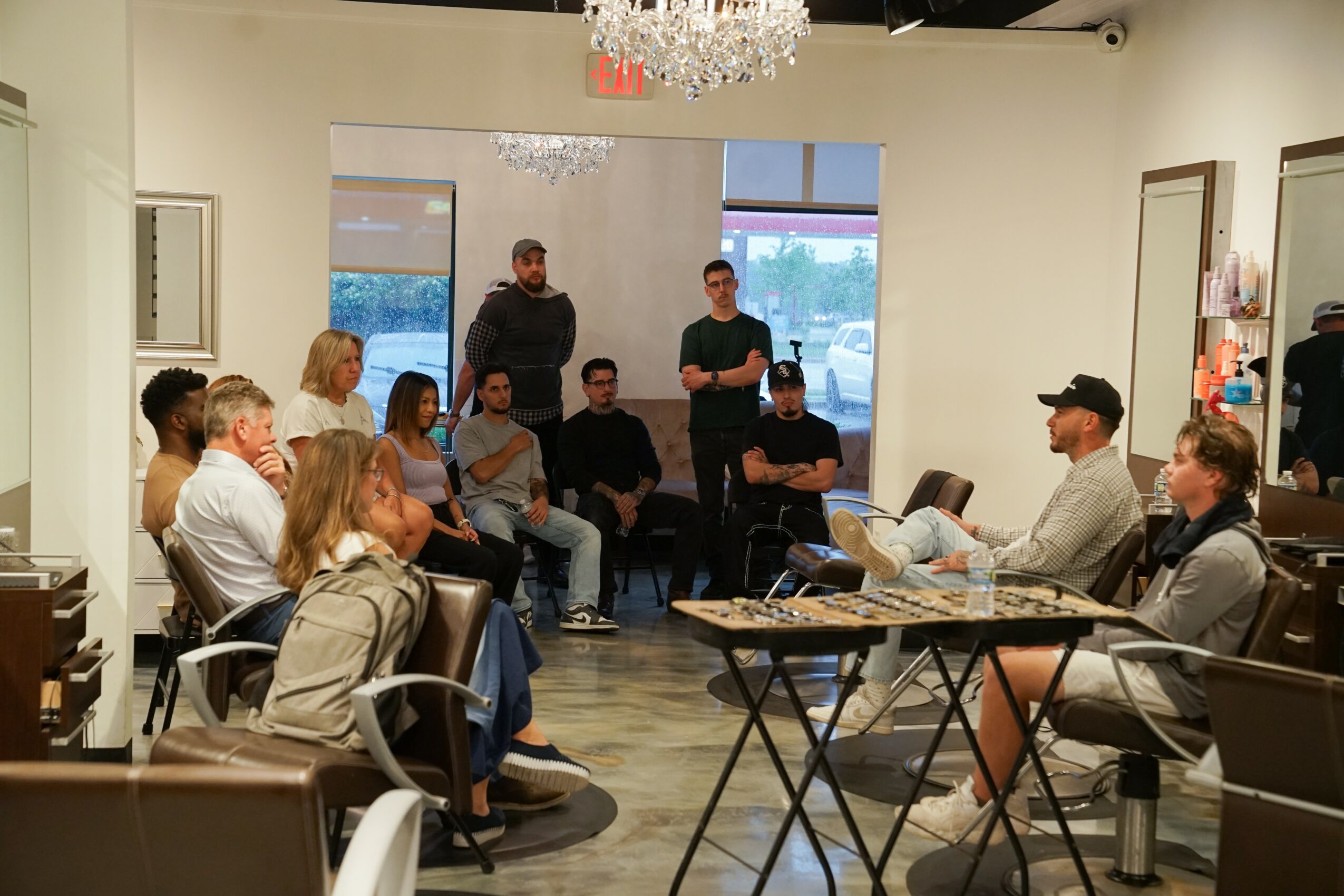 Group of people seated and standing in a room with white walls, chandelier, and large windows, engaged in discussion.