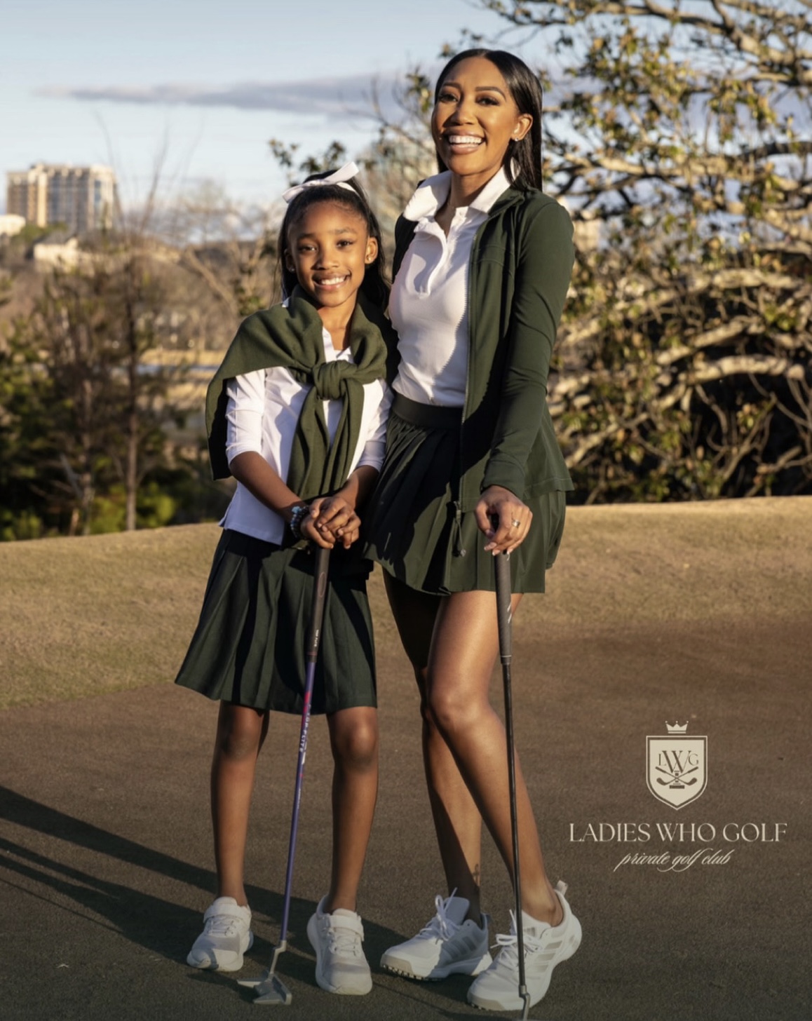 Two smiling women in golf attire holding golf clubs outdoors with trees and buildings in background.