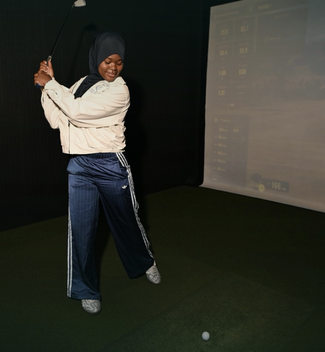 Person in hijab holding a golf club indoors, standing on green carpet, with a large screen in the background.