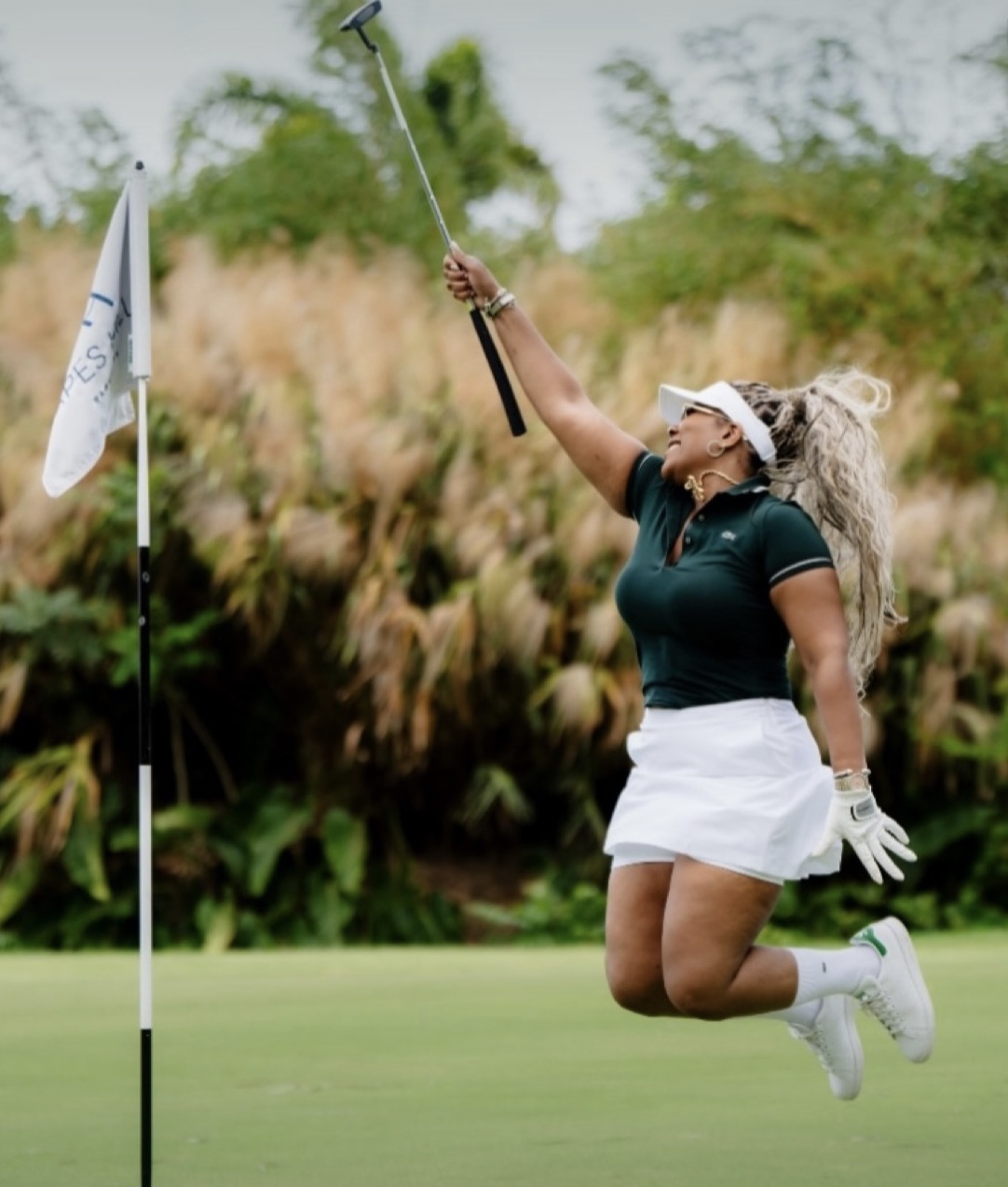 Woman jumping on golf course, holding golf club, near flagstick, with trees and bushes in background.