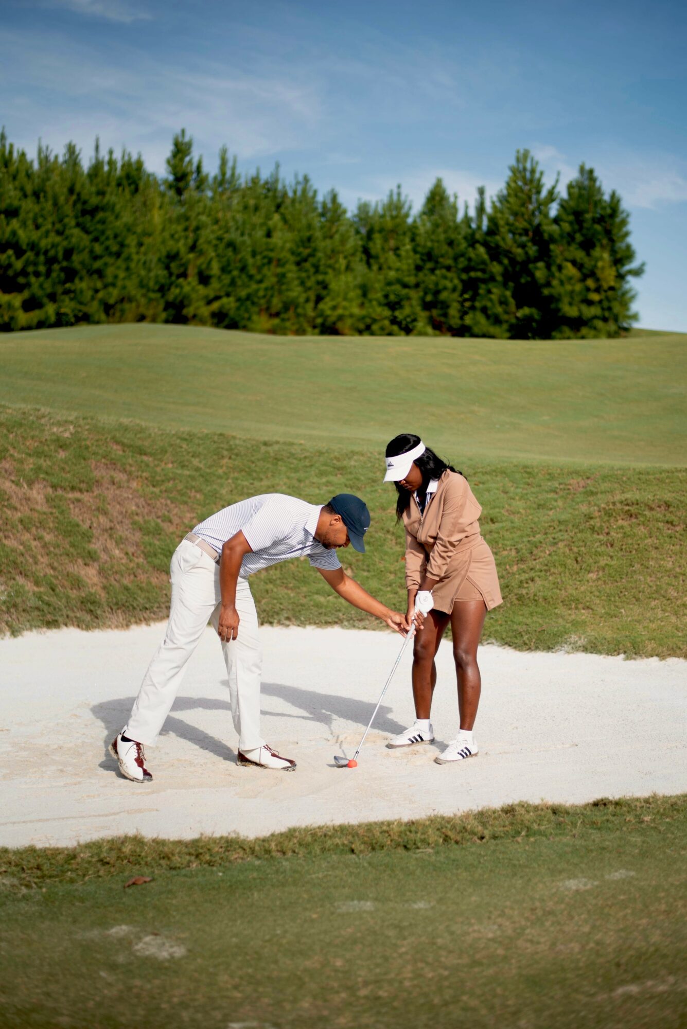 Two people on a golf course, one helping the other with a golf club in a sand trap, trees and sky in background.