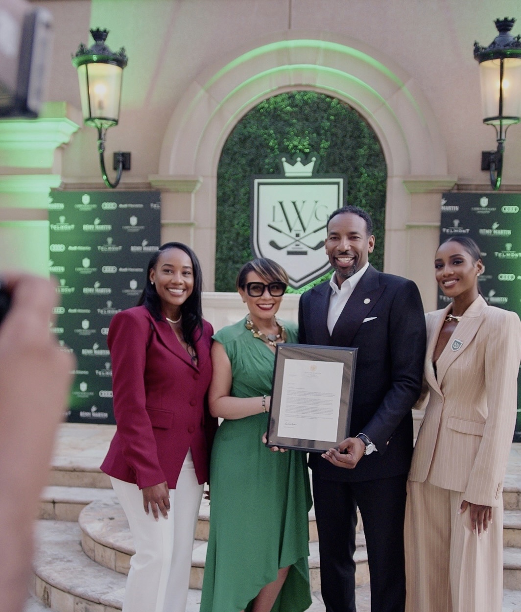 Four people standing outdoors in front of an arched structure with greenery, holding a framed document, smiling.