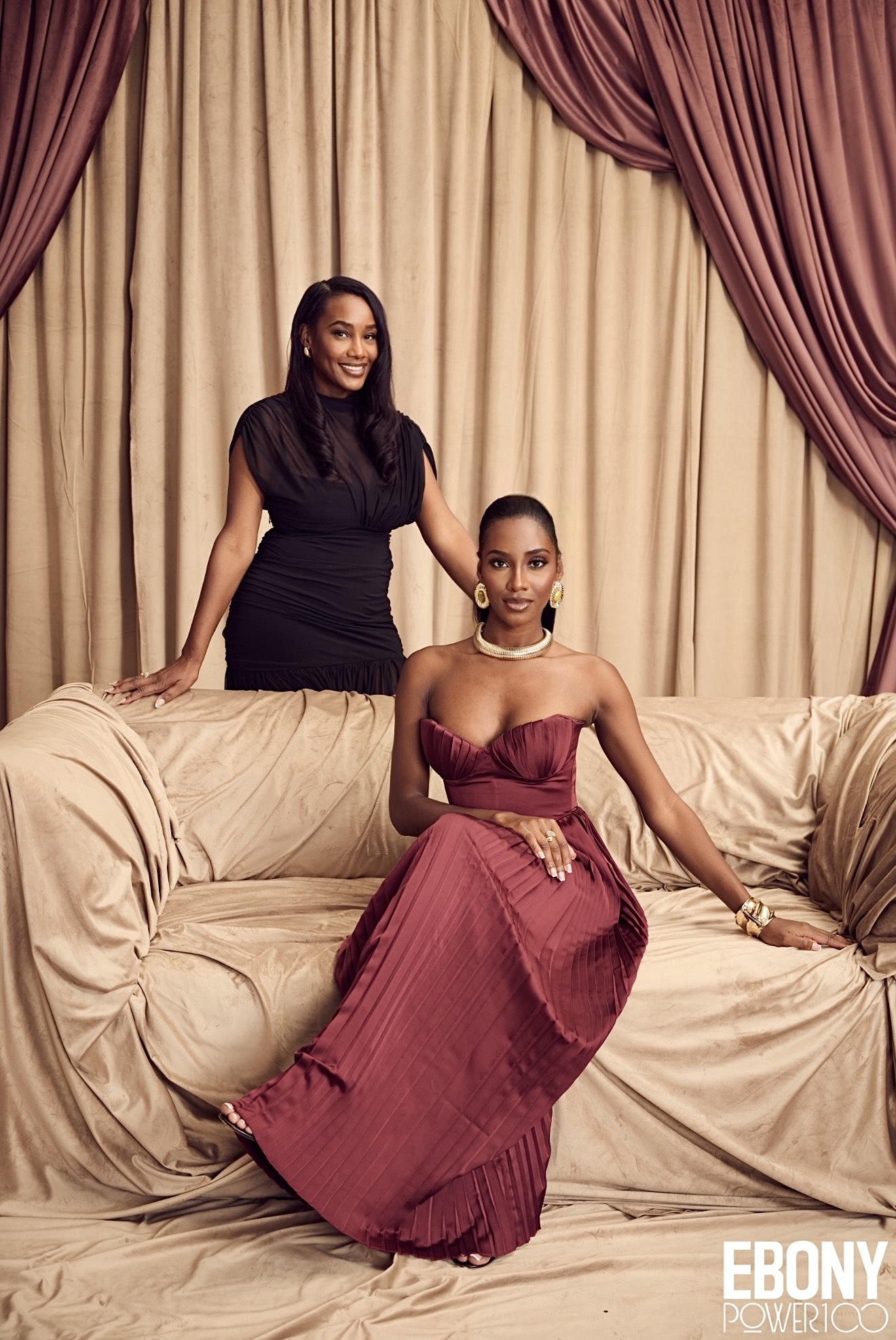Two women in elegant dresses pose on a beige sofa with curtains in the background. One stands, the other sits, both smiling.