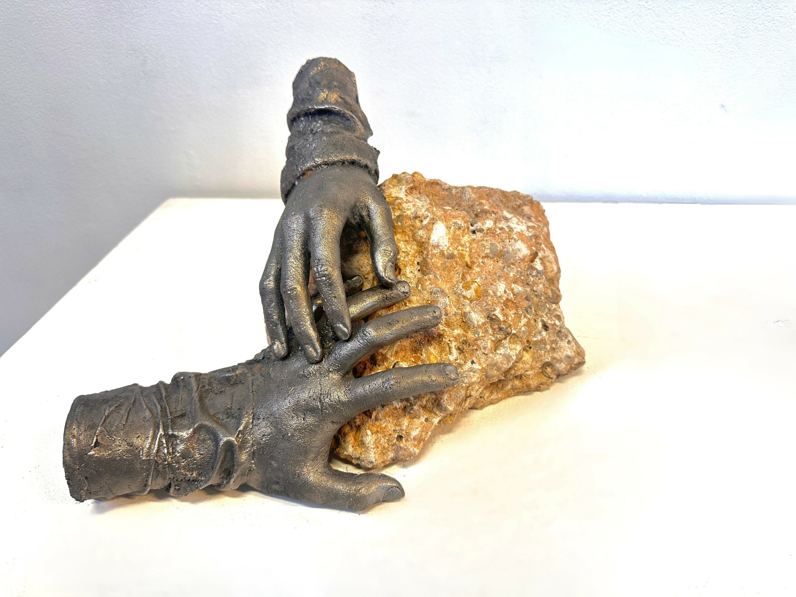 Two metallic hands, one holding a rock, are positioned on a white surface. The hands appear to be sculptures.
