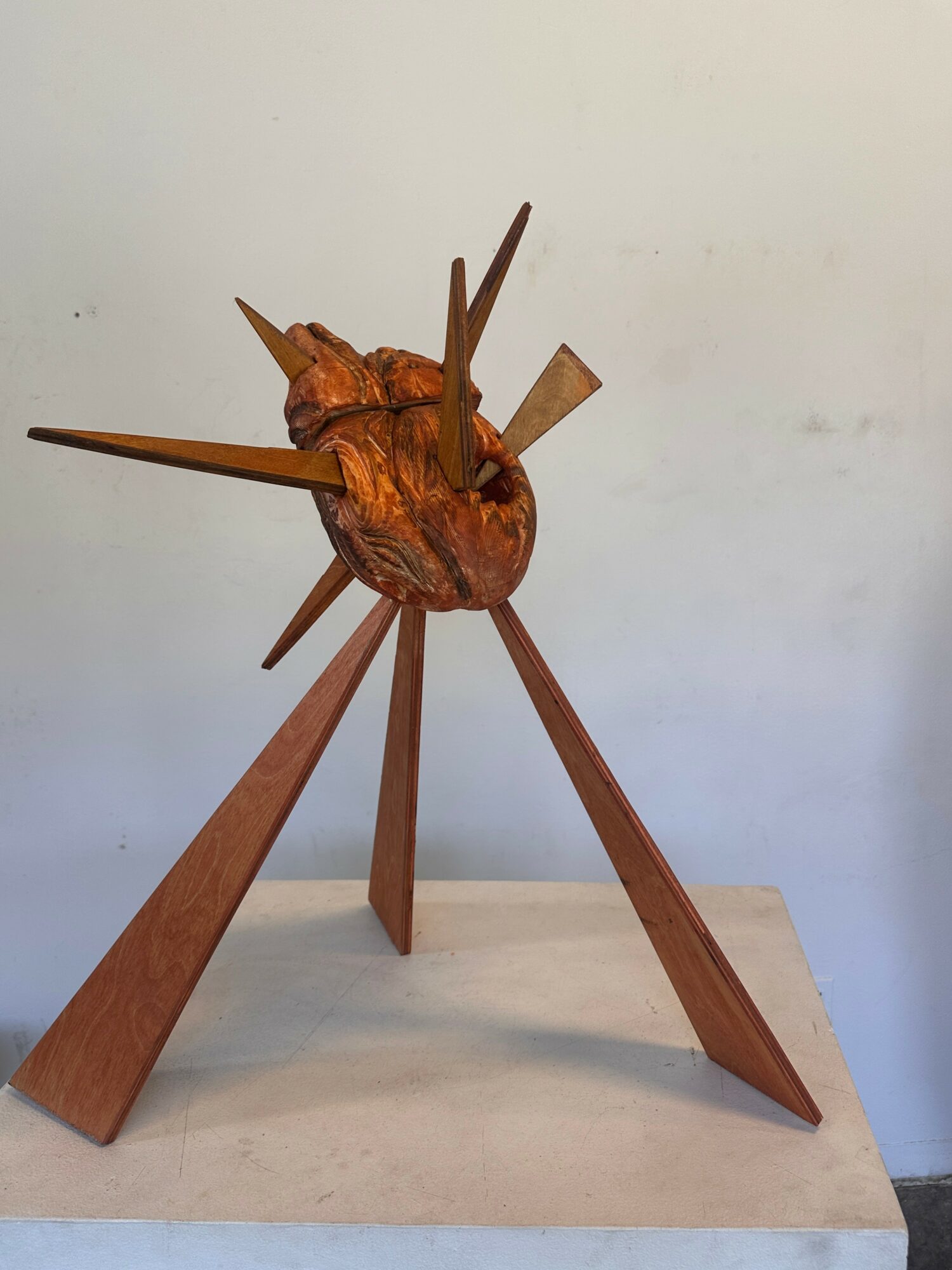 Wooden sculpture of a heart with pointed wooden spikes protruding in various directions, mounted on a wooden base.