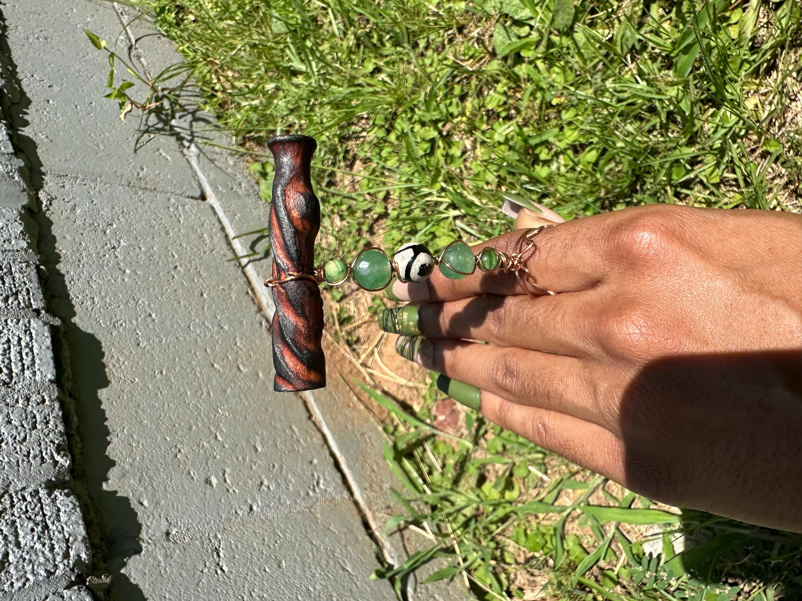 Person's hand holding a beaded bracelet with a wooden handle on grass and concrete surface, sunlight casting shadow.