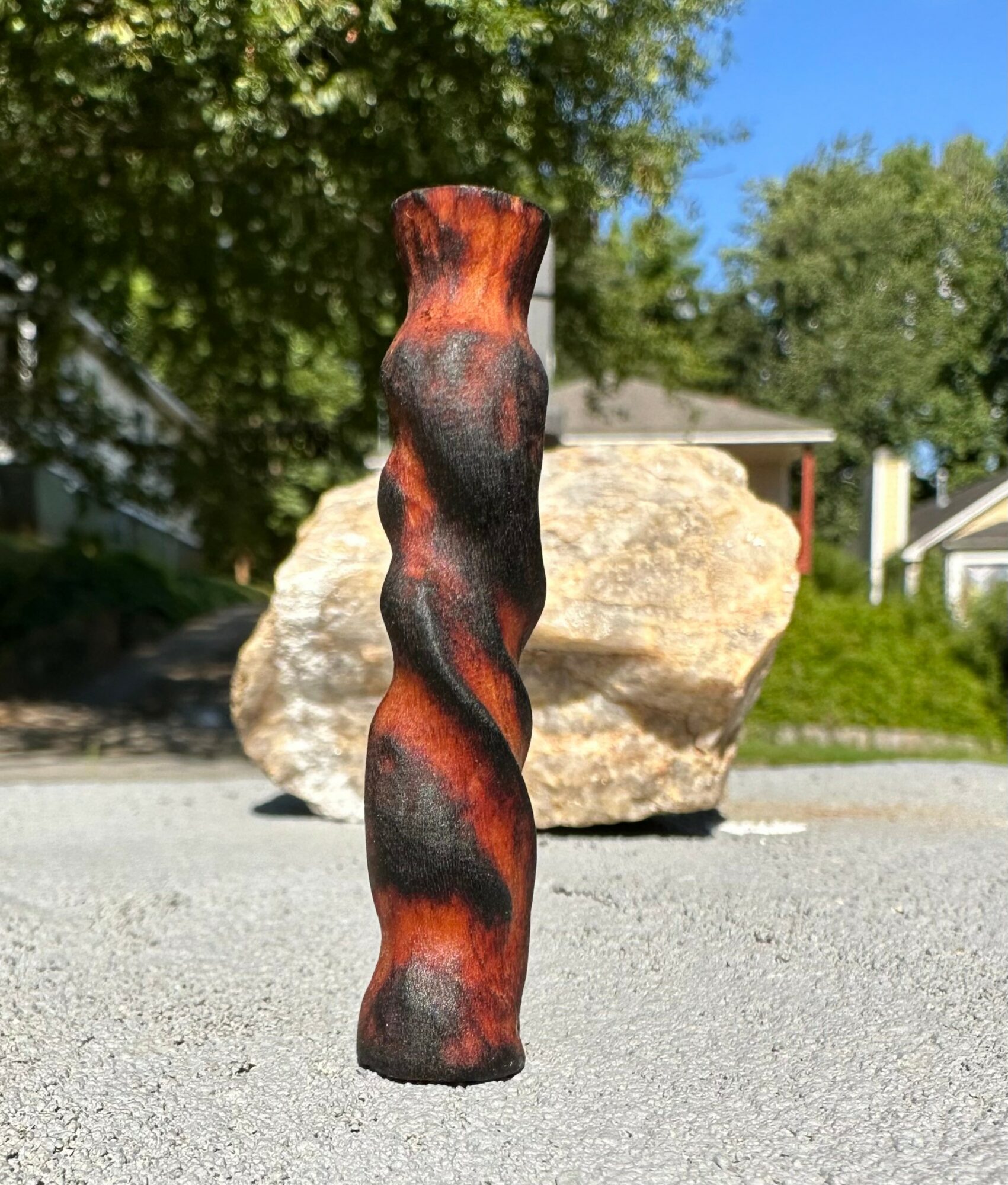 A twisted, reddish-brown wooden sculpture stands outdoors with a large rock and trees in the background.