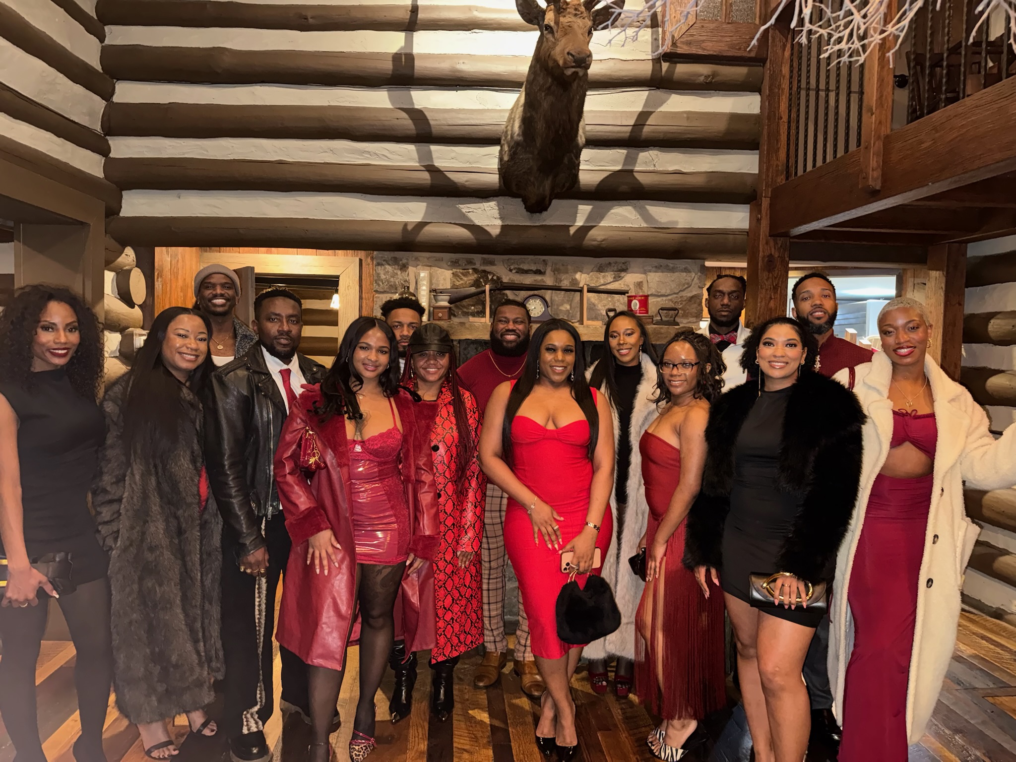 Group of people standing indoors, some wearing red dresses, in a log cabin-style room with a mounted animal head and basketball hoop.