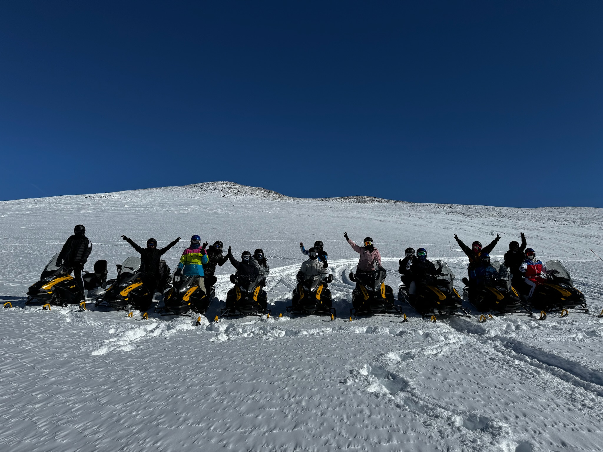 Group of people on snowmobiles in snowy landscape with a clear blue sky, some raising their arms.