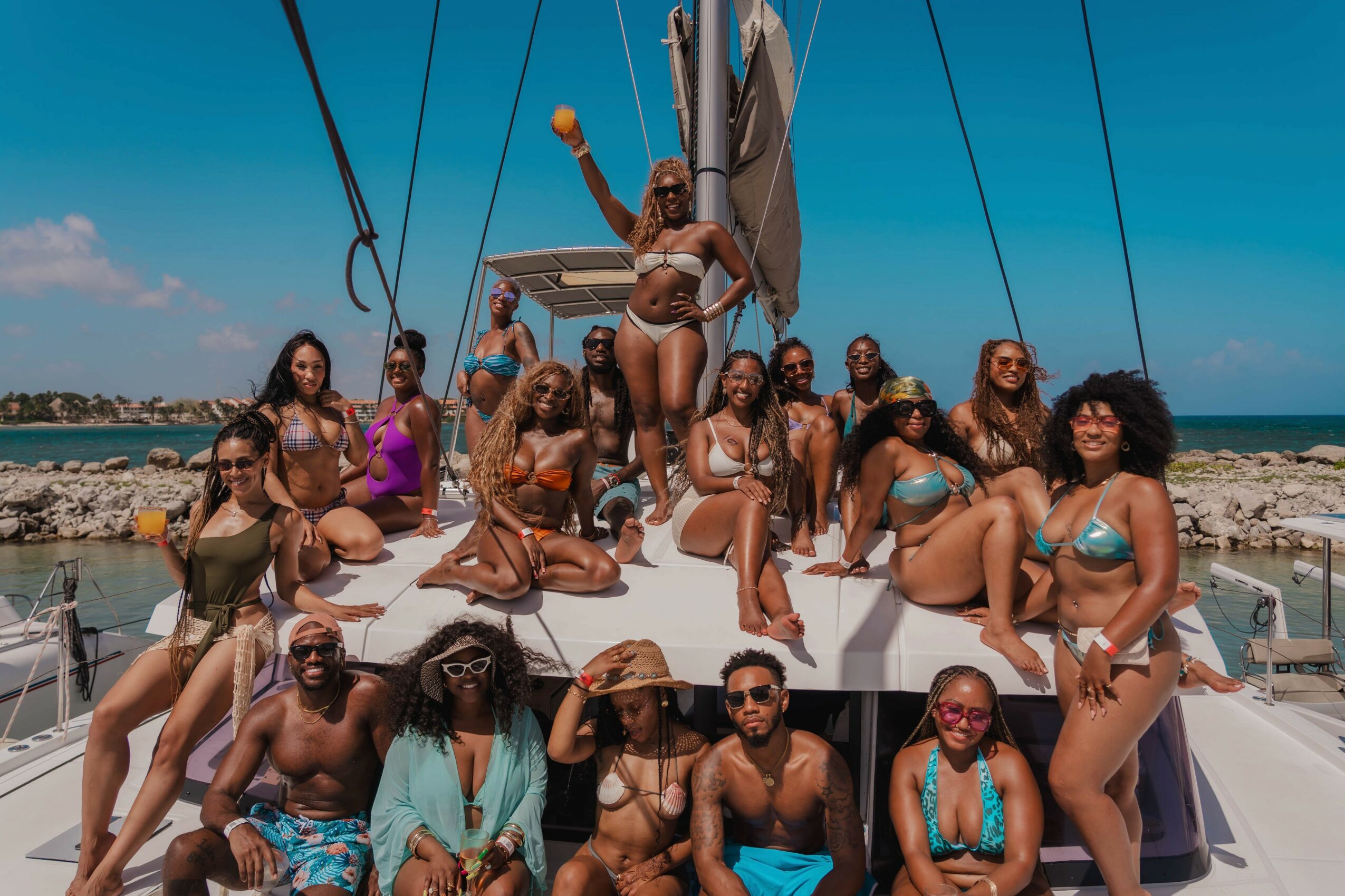 Group of women in swimsuits on a boat, some sitting and some standing, with a woman at the top holding a towel, on a sunny day by the water.