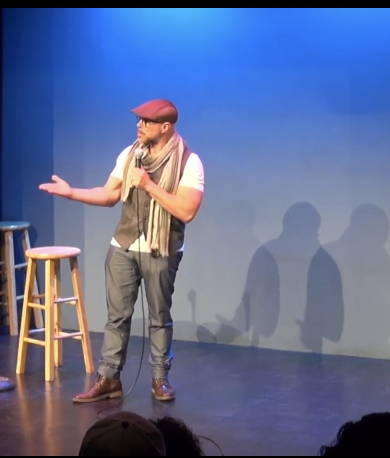 Person standing on stage with two stools, gesturing with one hand, shadows cast on blue background, audience visible at bottom.