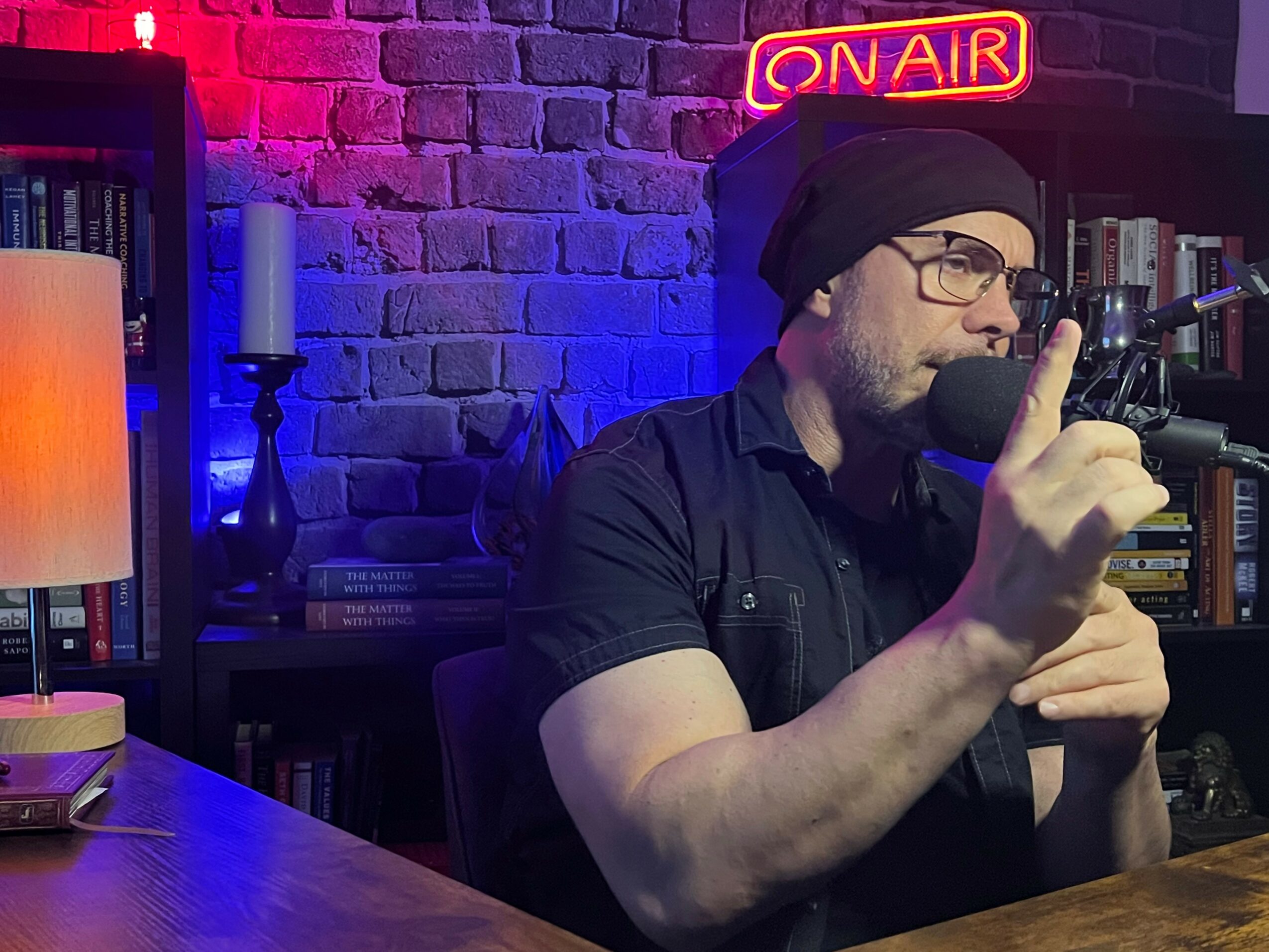 Man wearing glasses and a beanie speaking into a microphone in a studio with bookshelves and a neon 'ON AIR' sign