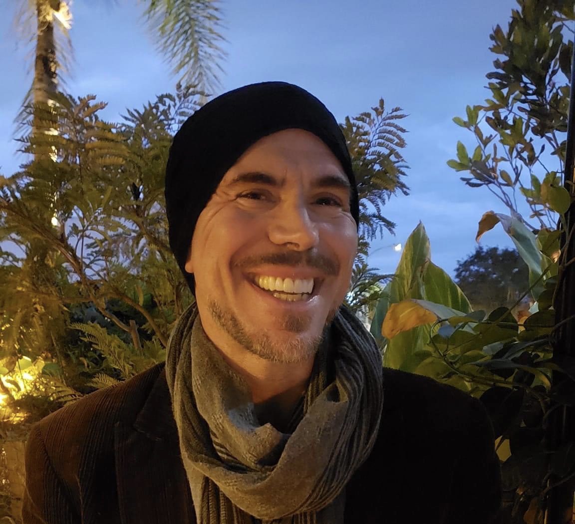 Smiling man wearing a black beanie and scarf outdoors with trees and blue sky in background.