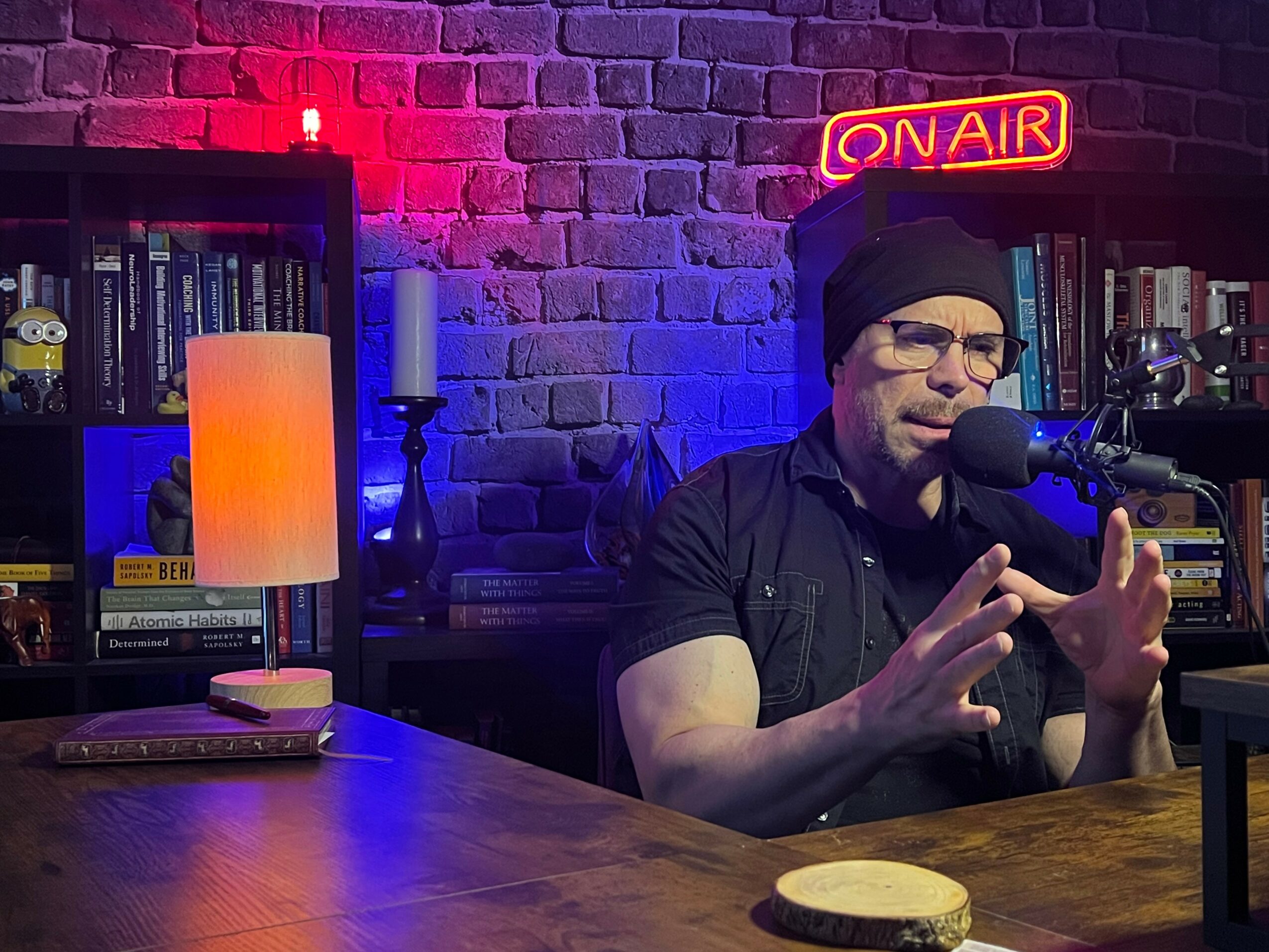 Person wearing glasses and a beanie speaking into a microphone in a studio with bookshelves and a neon 'ON AIR' sign