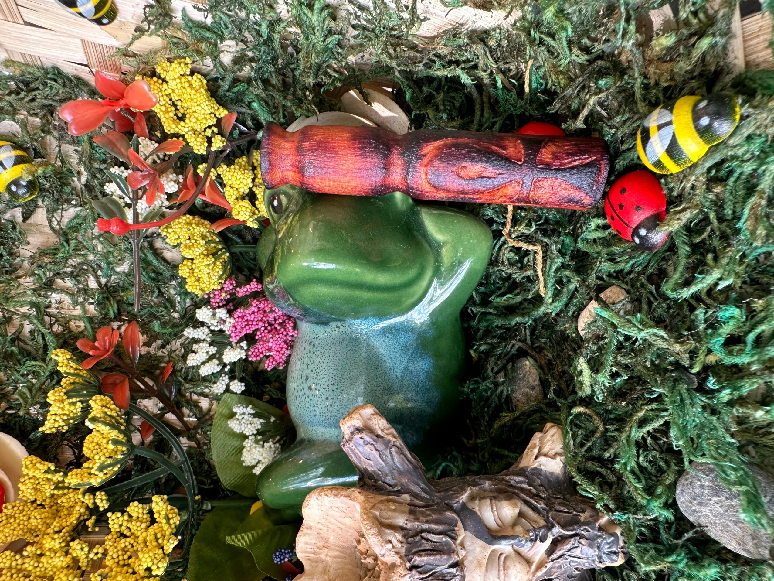 Colorful garden scene with a frog figurine, flowers, a ladybug, and a bee among greenery.