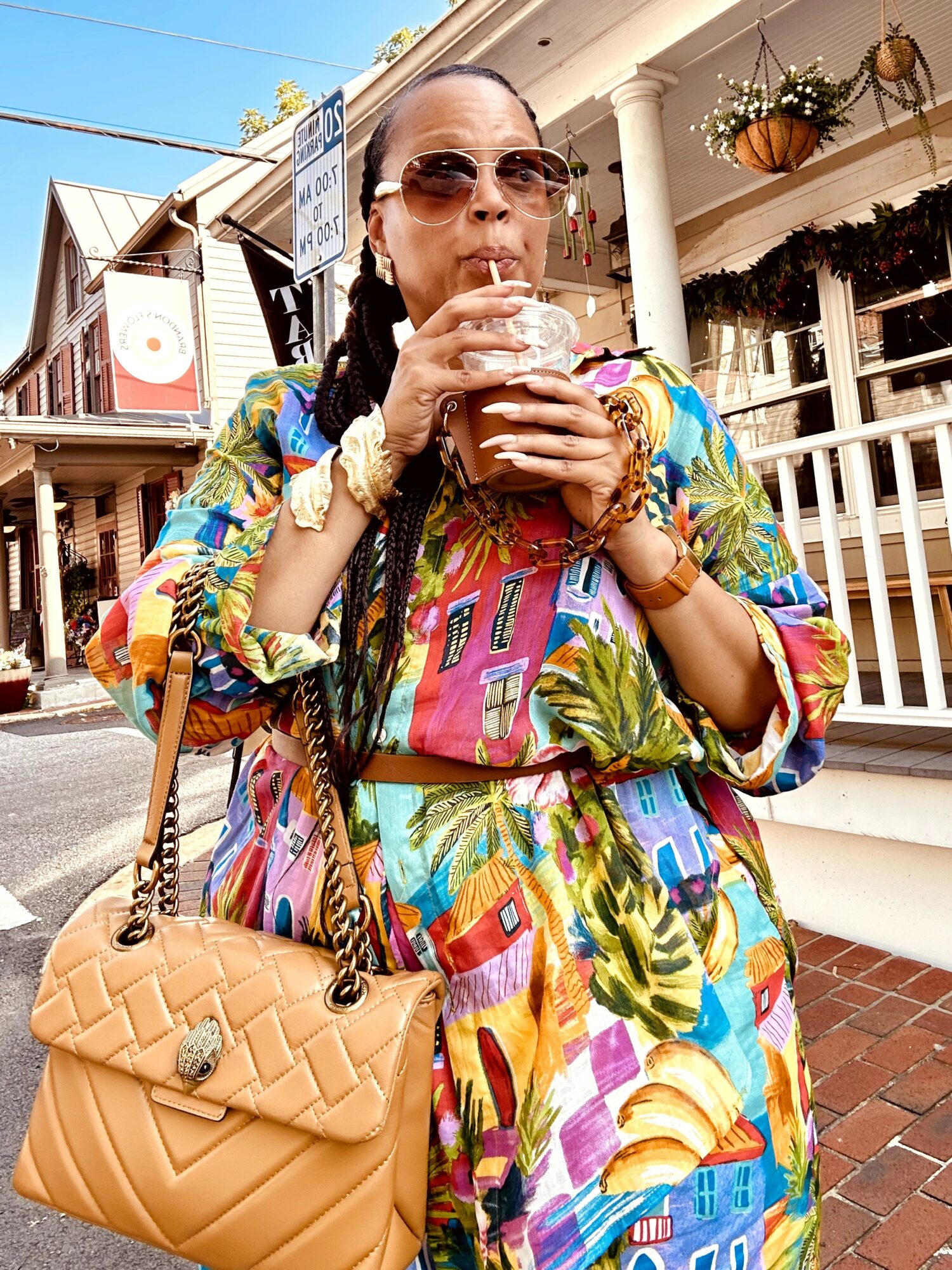 Woman in colorful dress drinking from a glass outdoors, wearing sunglasses, with buildings and hanging plants in background.