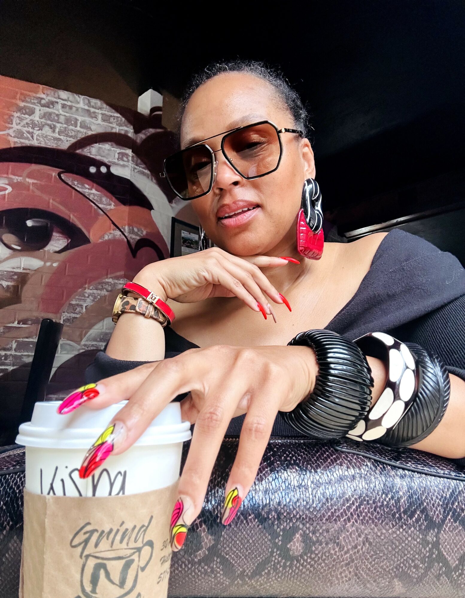 Woman with sunglasses and colorful earrings sitting at a table, holding a coffee cup, with a brick wall background.