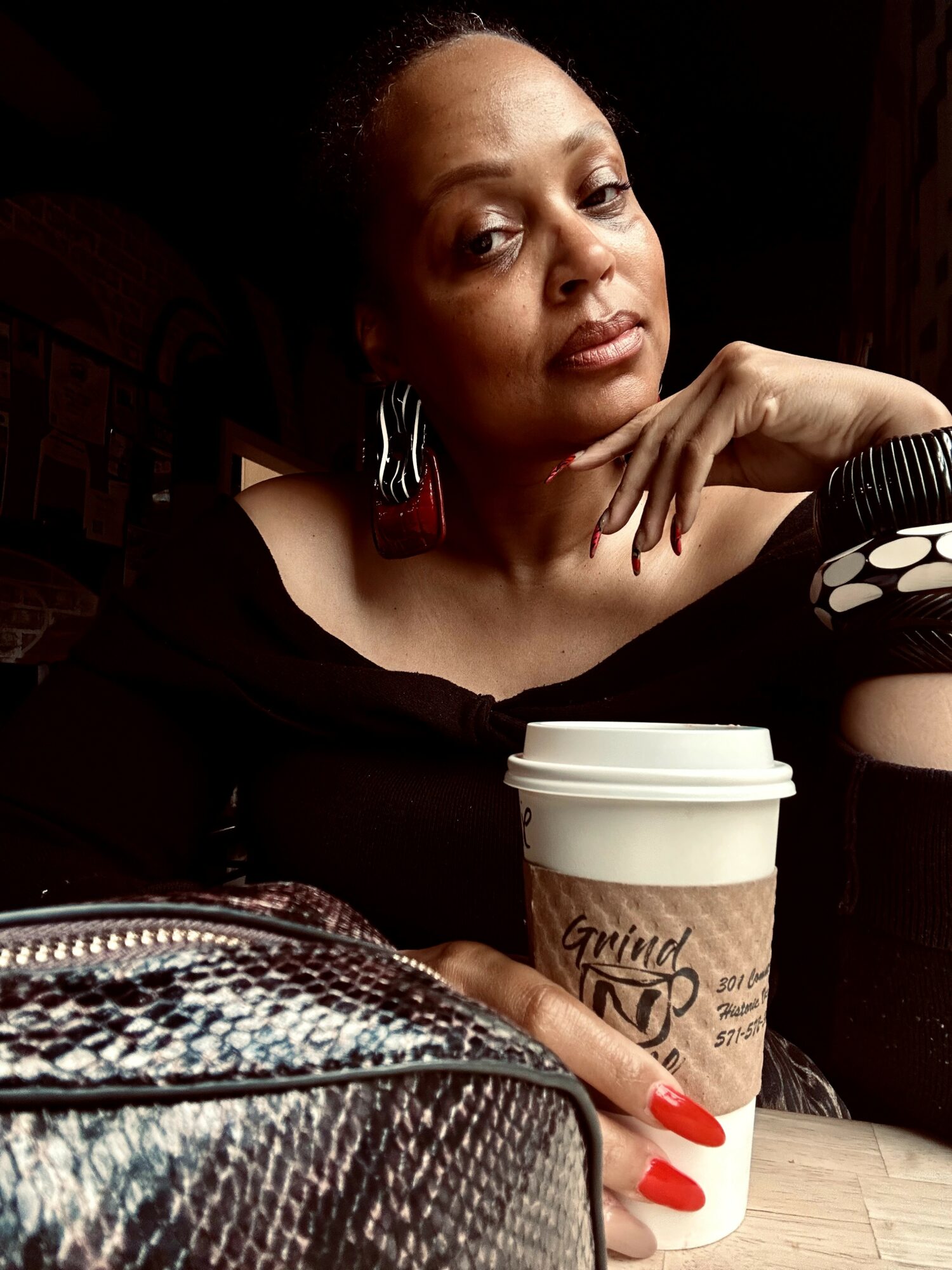 Woman with dark hair and earrings resting her chin on her hand, holding a coffee cup, with a bag in front.