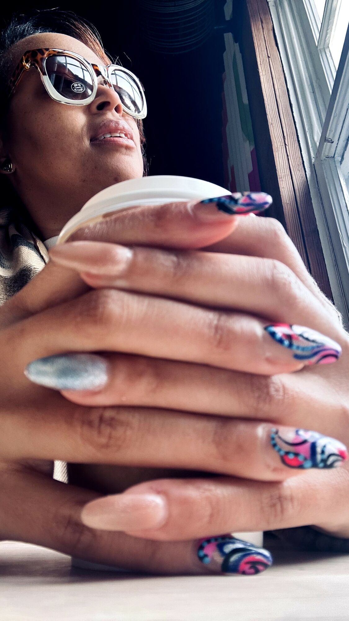 Woman with sunglasses looking out a window, holding a cup, with decorated nails, close-up of hands and face.