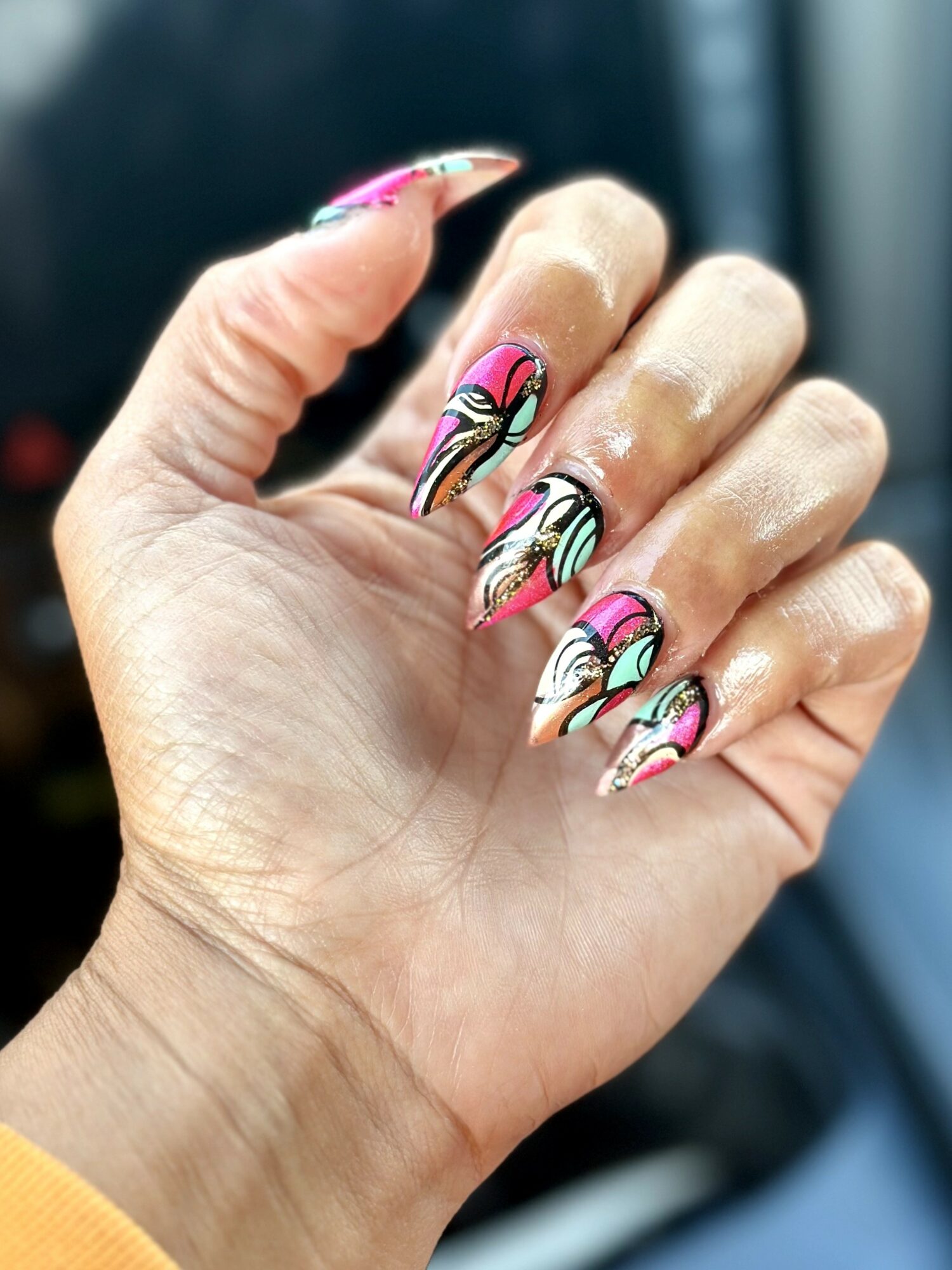 Hand with decorated nails featuring pink, black, white, and gold swirl patterns, held against a blurred background.
