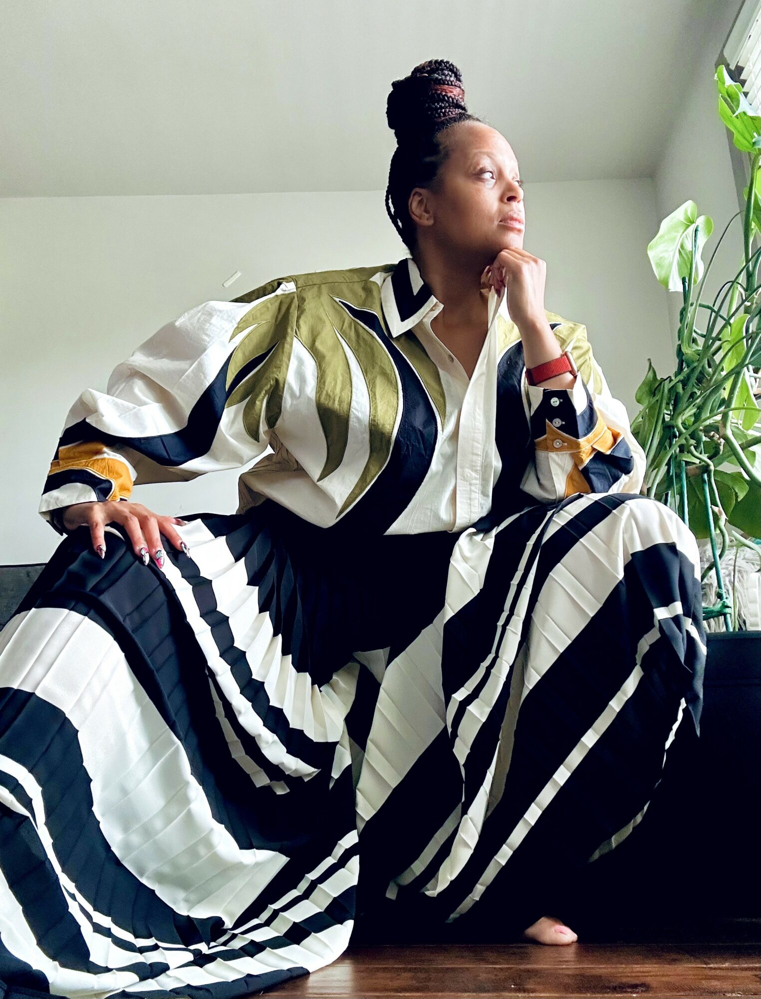 Woman sitting on a dark surface near a window with green plants, wearing a striped outfit, looking thoughtfully outside.