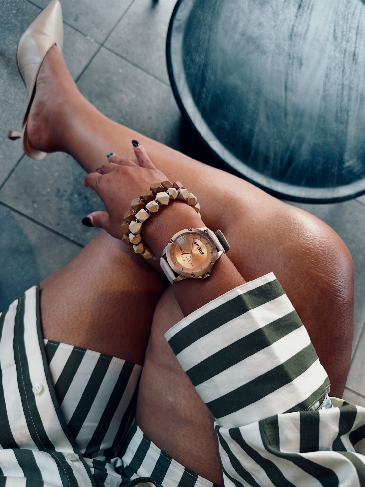 Person sitting with crossed legs, wearing striped clothing, white high heel, and jewelry, near a round table on tiled floor.