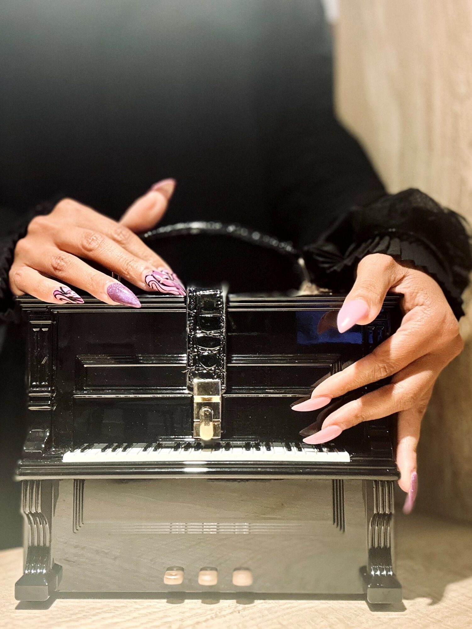 Person's hands playing a small black piano, with painted nails, on a wooden surface.