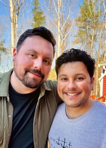 Two men smiling outdoors with trees and a red building in the background.
