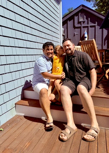Three people sitting on a wooden deck outside a house, smiling and hugging, with a blue house wall behind them.