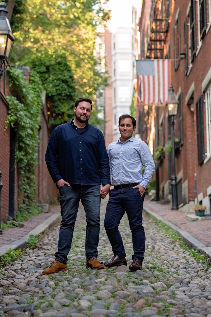 Two men stand on a cobblestone street lined with brick buildings and greenery, holding hands and smiling.