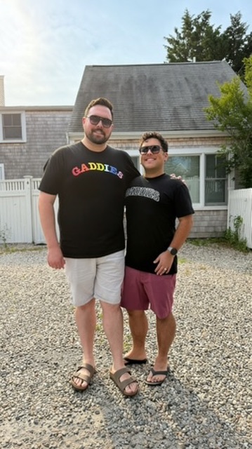 Two men standing outdoors on gravel, one taller with a beard and sunglasses, the other shorter with sunglasses, in front of a house.