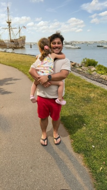 Person holding a child near a waterfront with a ship and boats in the background, smiling, on a sunny day.