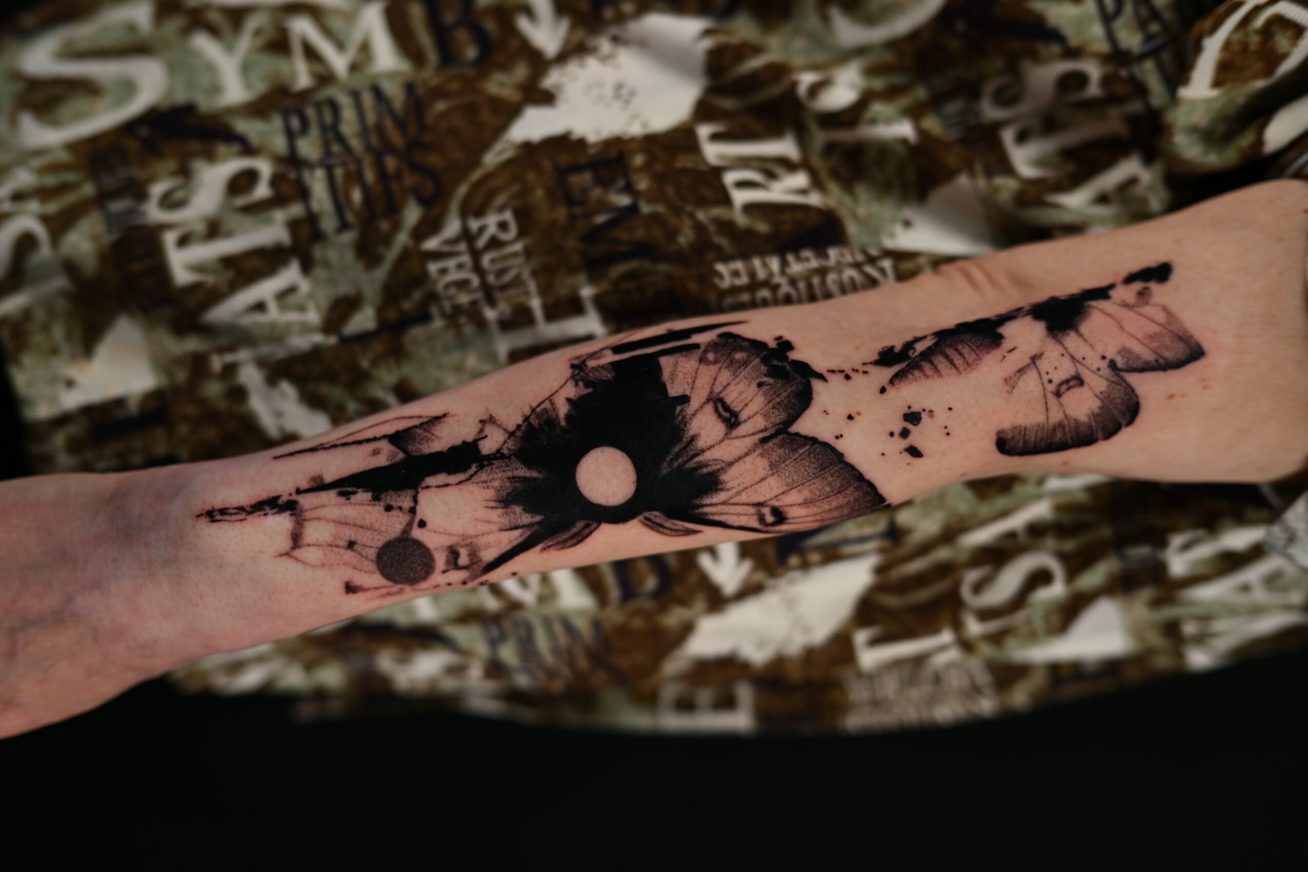 Forearm with a black butterfly tattoo and abstract ink splashes, background with patterned fabric, skin tone visible.