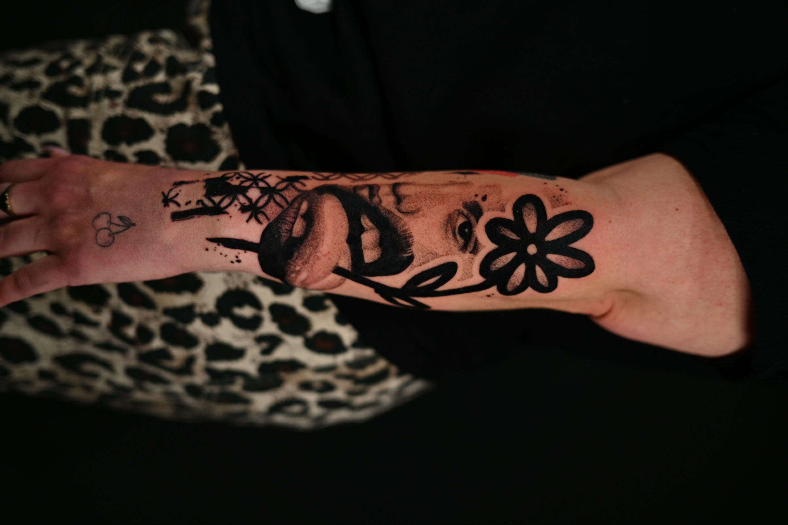 Forearm tattoo of a woman's face, lips, and a flower, with a leopard print fabric in the background.