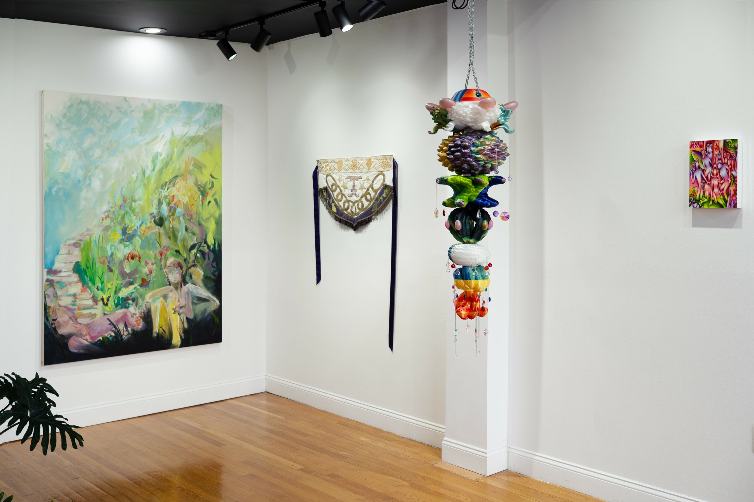Art gallery with colorful paintings and hanging sculpture, wooden floor, white walls, and ceiling spotlights.