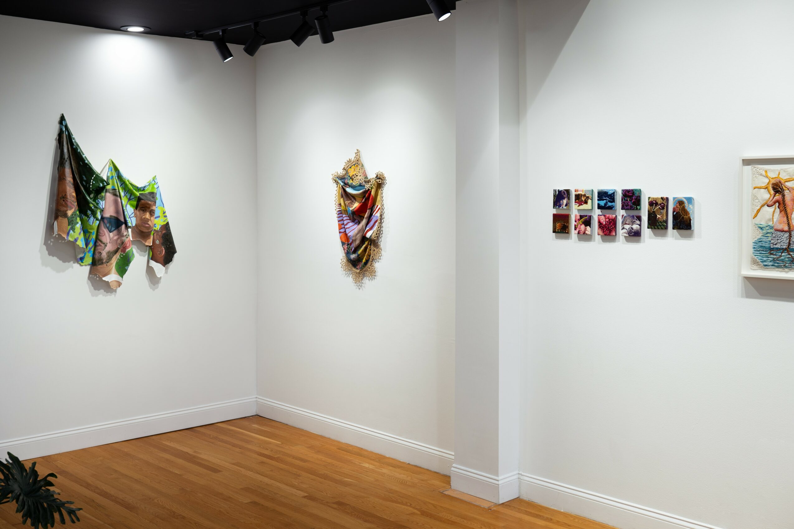 Art gallery with colorful fabric sculptures and small framed artworks on white walls, wooden floor, and ceiling lights.