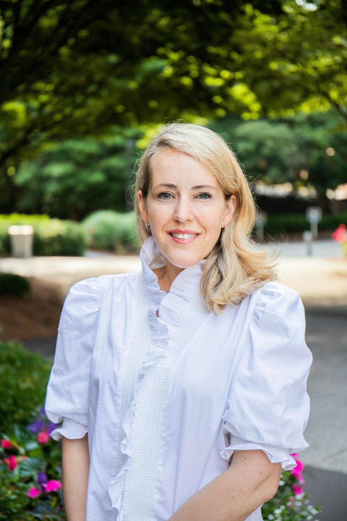 Smiling woman with blonde hair in a white blouse outdoors with trees and flowers in background.