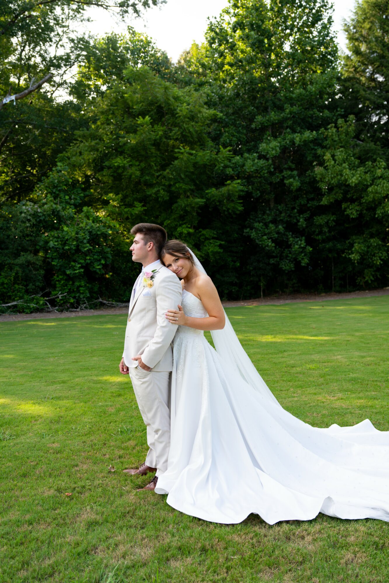 Bride in a white wedding dress leaning on groom in a light-colored suit outdoors, green trees in background