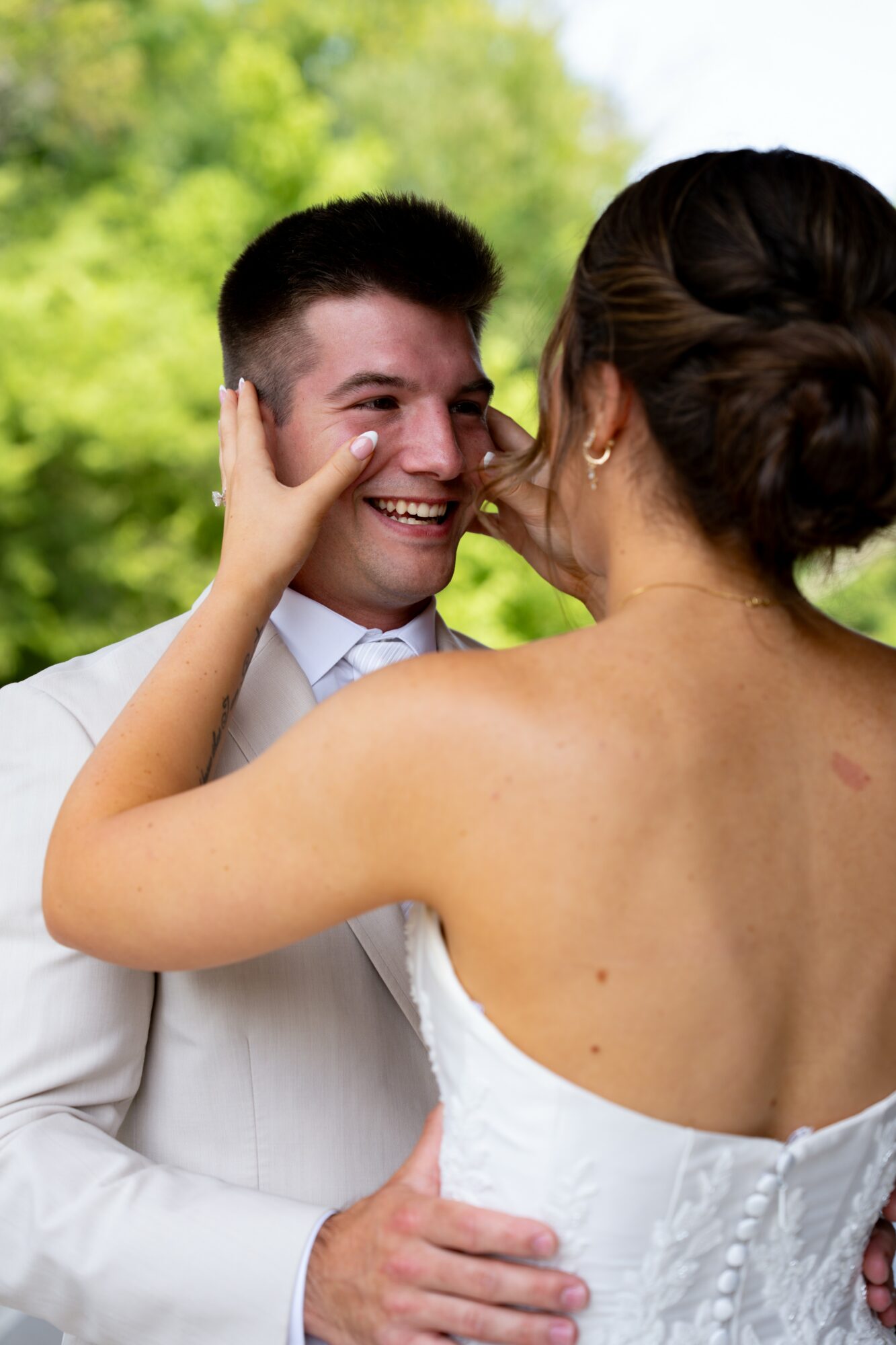 A woman and a man are close, smiling, with the woman touching the man's face outdoors, greenery in background.