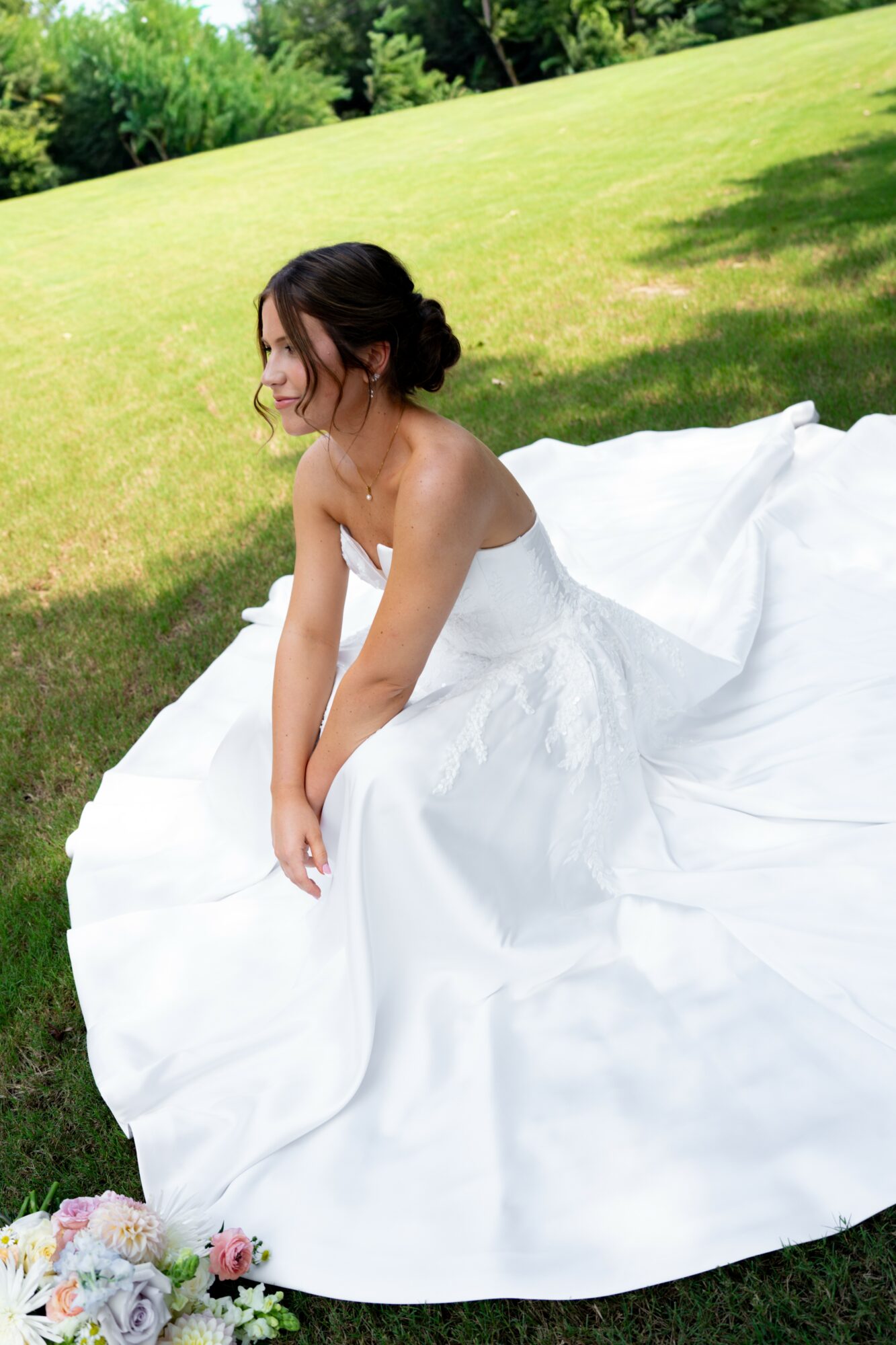 Woman in wedding dress sitting on grass in a park with trees in the background.