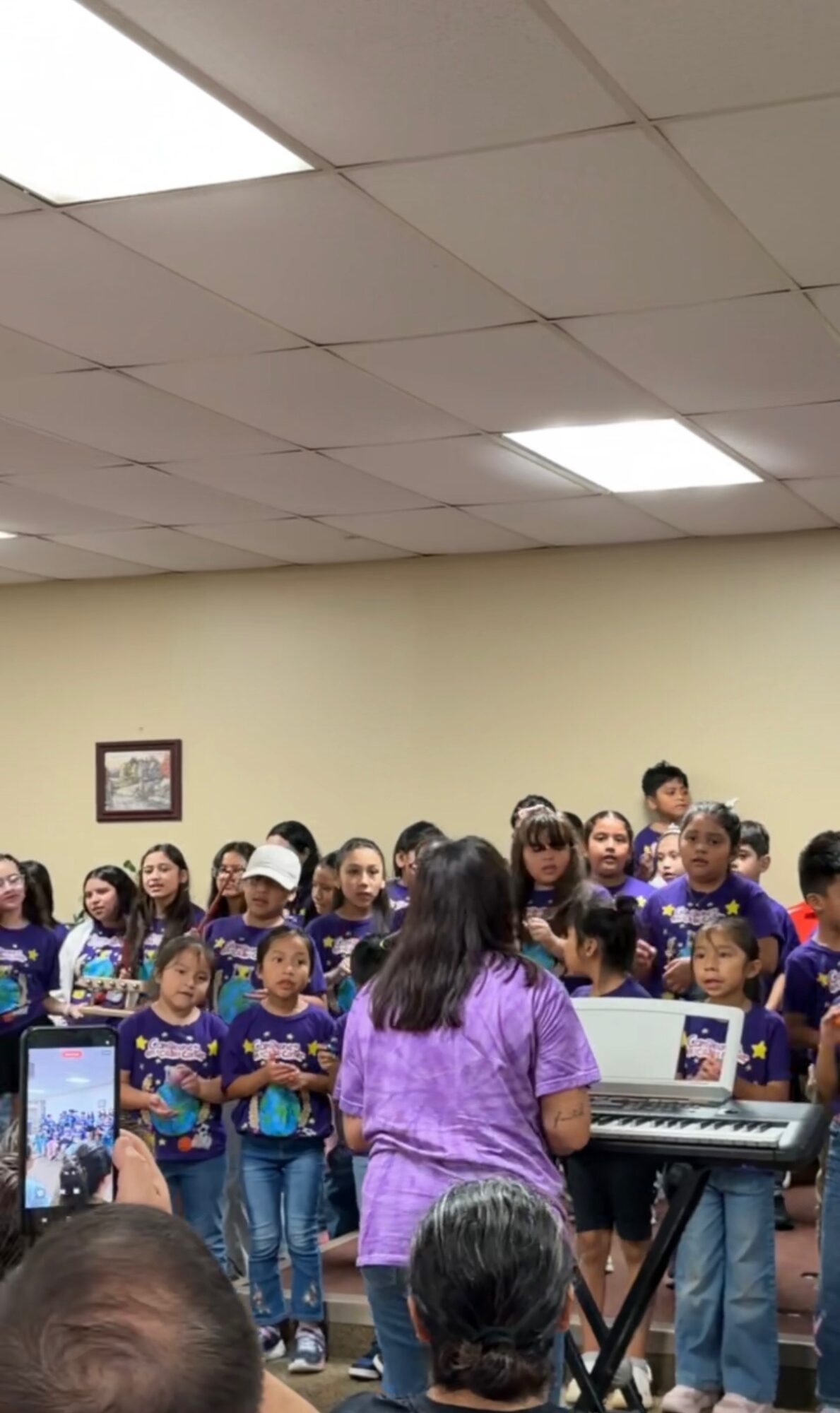 Group of children and a woman in purple shirt in a room with ceiling lights, some children holding papers, woman at keyboard.