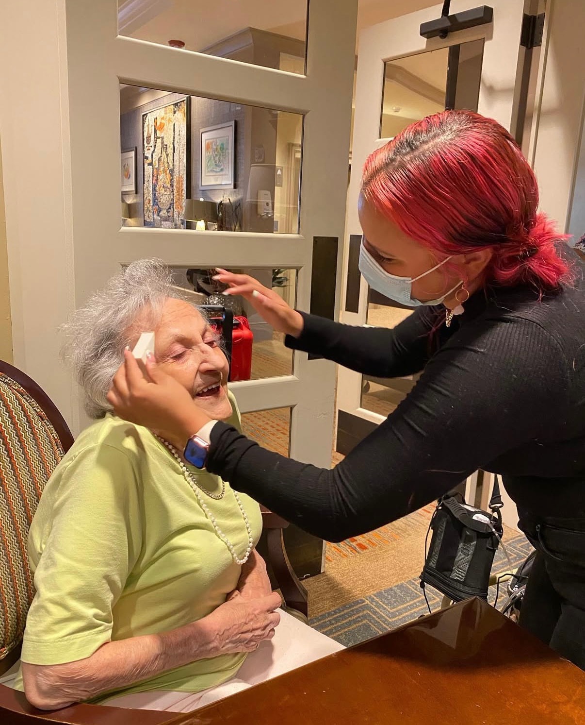 A caregiver with red hair and a face mask gently touches an elderly woman's face, who is smiling and sitting at a table.