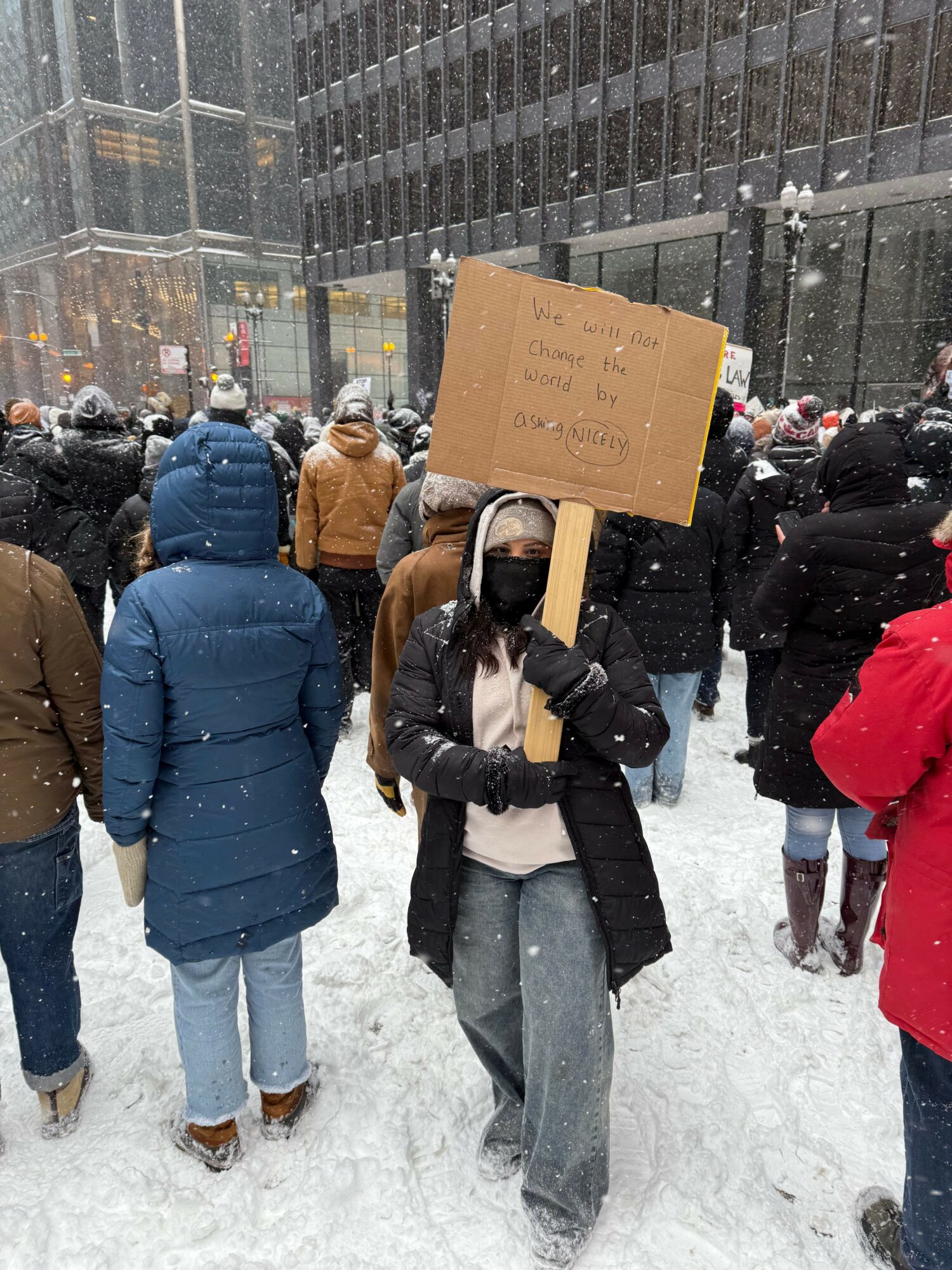 People gathered outdoors in snowy weather, one person holding a cardboard sign, others wearing winter clothing, urban background.