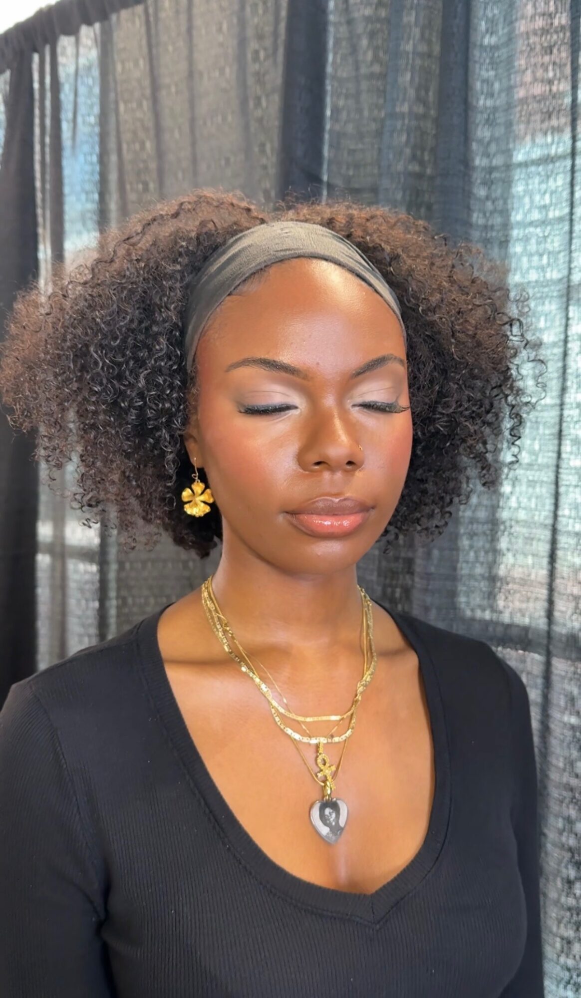 Woman with curly hair, wearing a headband, gold jewelry, and a black top, with eyes closed, in front of curtains.