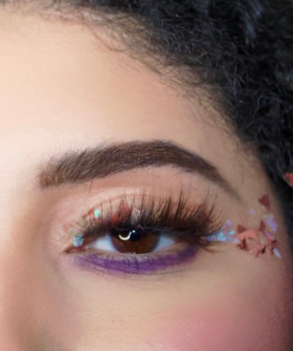 Close-up of a person's eye with makeup, glitter, and decorative elements near the outer corner, with purple eyeliner and long eyelashes.