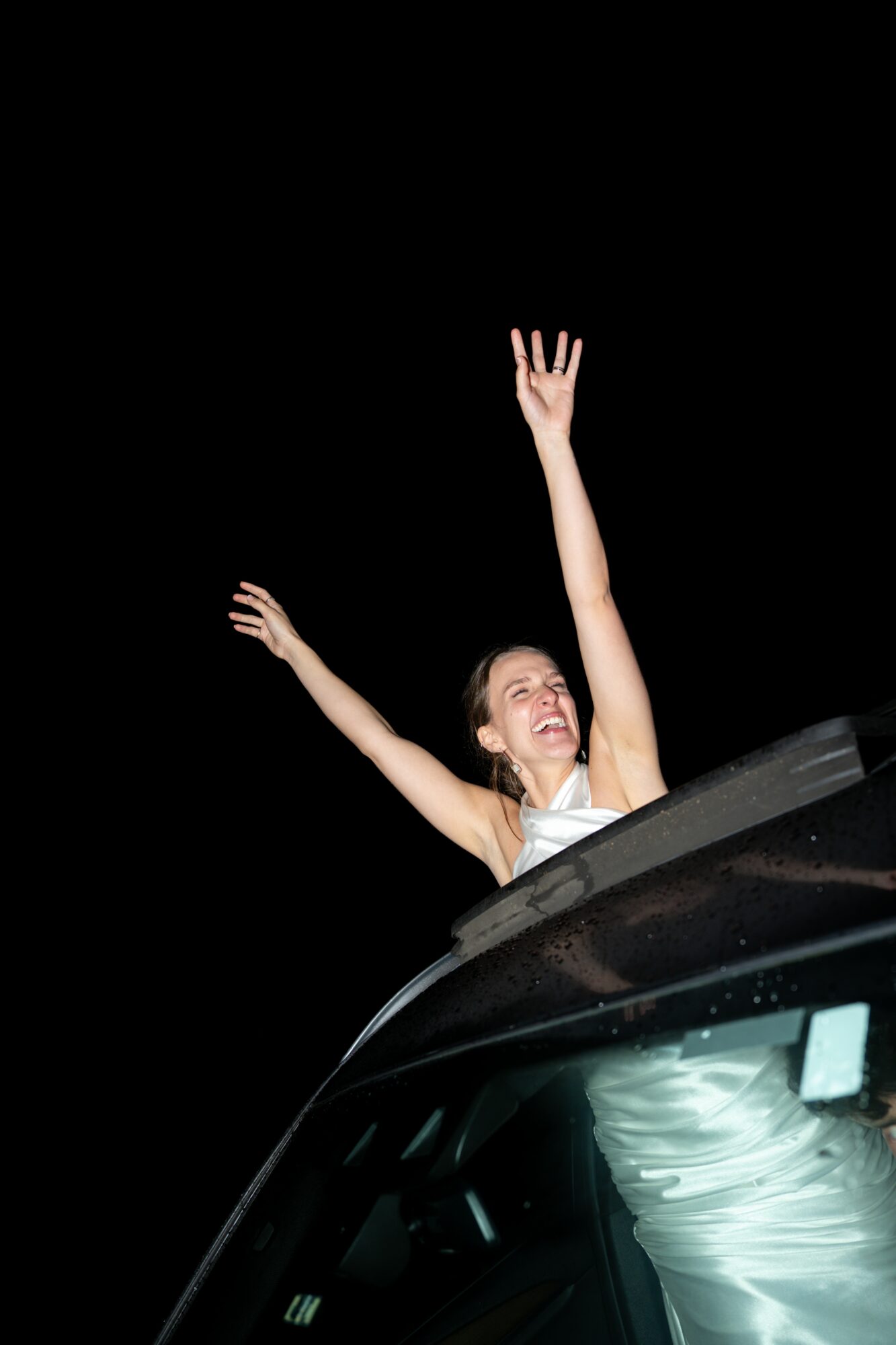 Person in a white dress with arms raised, smiling, leaning out of a car window at night.