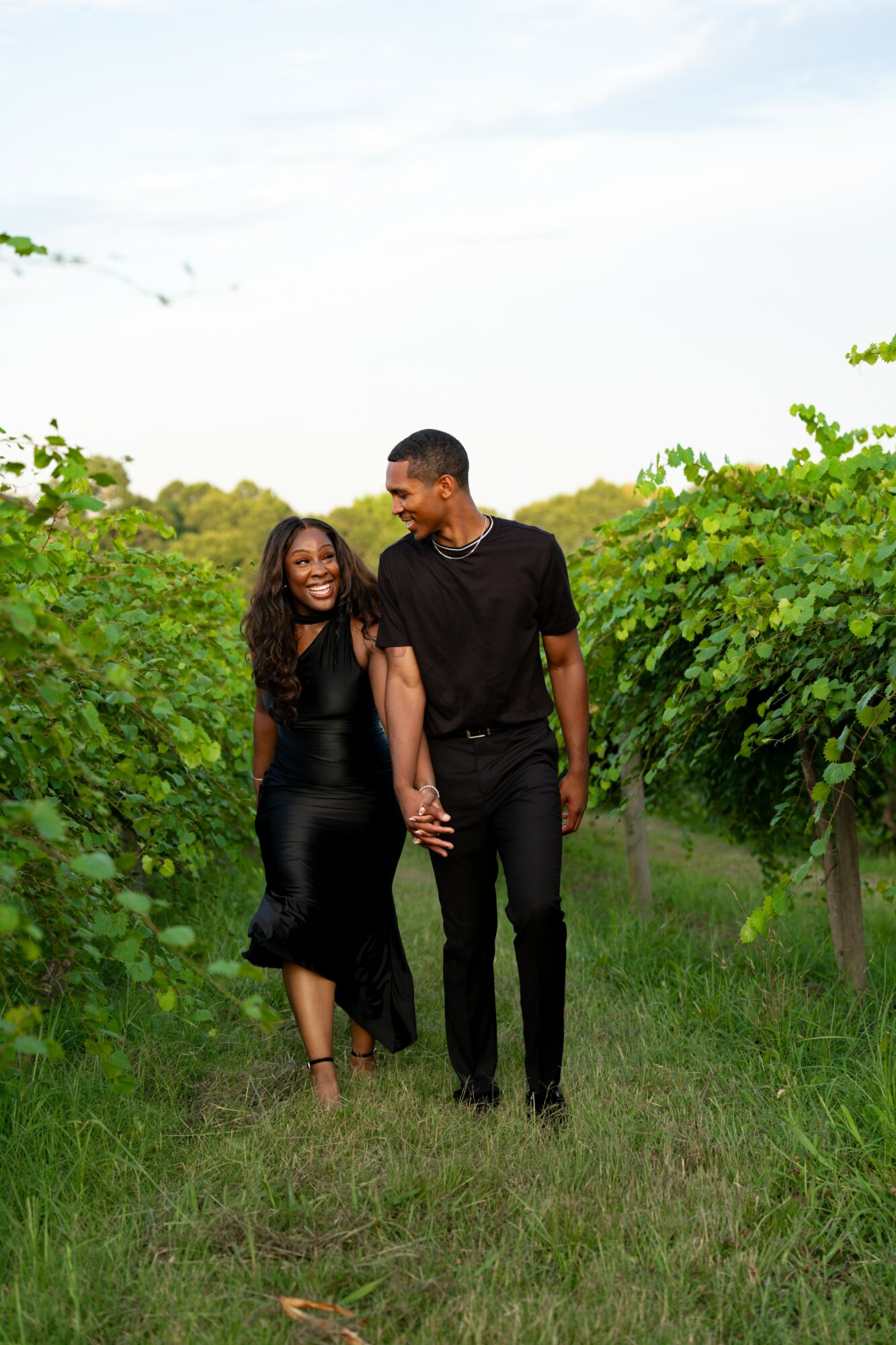 A smiling couple holding hands walking through a green vineyard, with trees and a blue sky in the background.