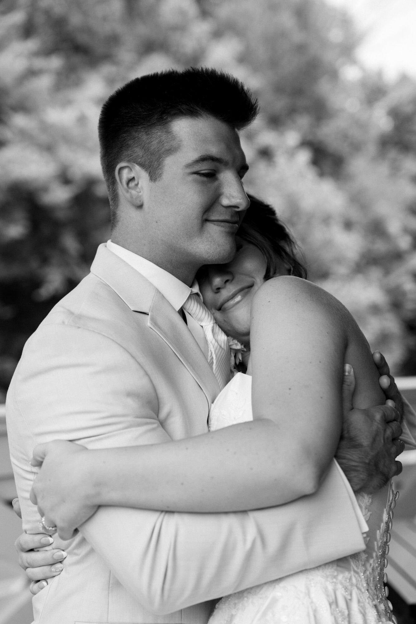 Two people hugging outdoors, one with short hair and the other with long hair, smiling and embracing each other.